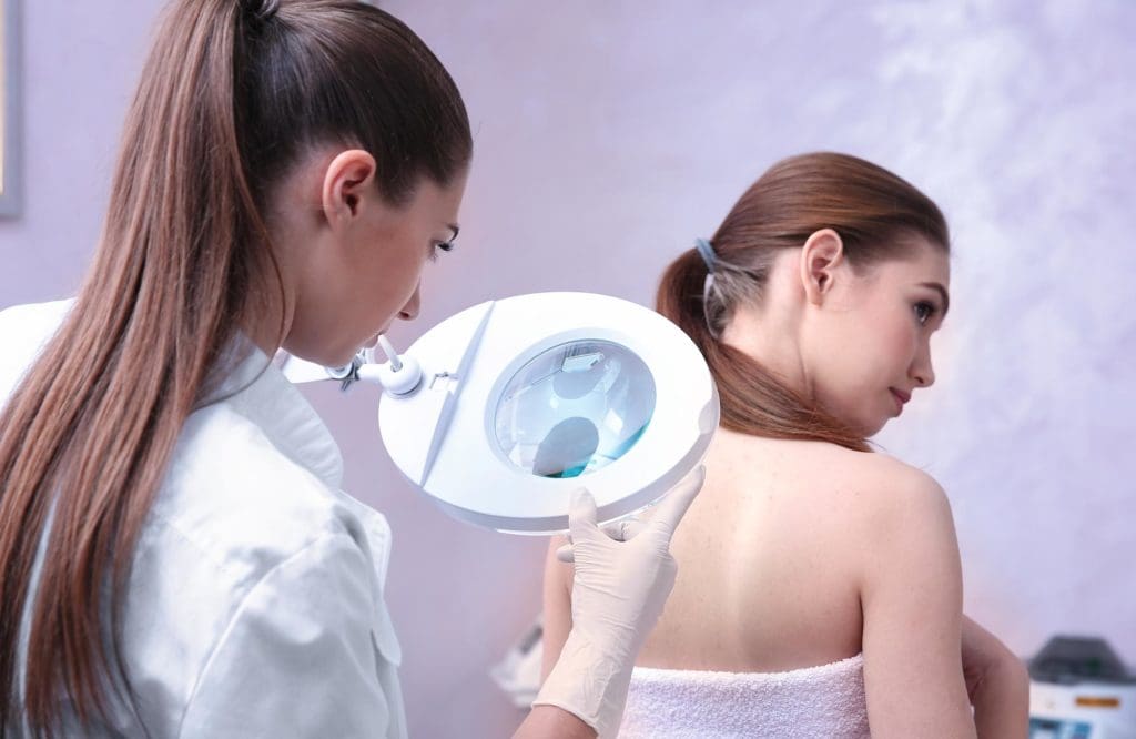 A doctor examines a patient's shoulder using a magnifying examination lamp during a skin check.